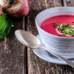 Recettes alcalines betterave: 5 recettes pour détoxifier votre corps 22 recettes alcalines betterave: Bol de velouté rose à la betterave garni de crème et ciboulette, posé sur une table en bois rustique avec une cuillère et des feuilles d’ail des ours.