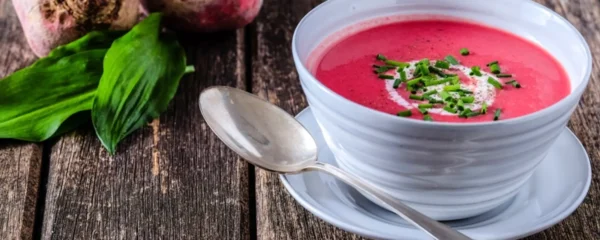 recettes alcalines betterave: Bol de velouté rose à la betterave garni de crème et ciboulette, posé sur une table en bois rustique avec une cuillère et des feuilles d’ail des ours.