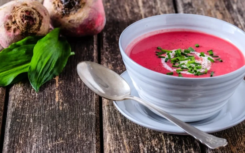 recettes alcalines betterave: Bol de velouté rose à la betterave garni de crème et ciboulette, posé sur une table en bois rustique avec une cuillère et des feuilles d’ail des ours.