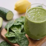 Verre de smoothie alcalin vert épais entouré de feuilles d'épinards, demi-avocat, concombre, citron et gingembre frais sur planche en bois, recette de smoothie alcalin maison