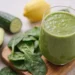 Verre de smoothie alcalin vert épais entouré de feuilles d'épinards, demi-avocat, concombre, citron et gingembre frais sur planche en bois, recette de smoothie alcalin maison
