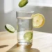 Verre d'eau avec rondelles de concombre et tranche de citron en lévitation autour du verre sur table en bois clair, recette d'eau alcaline maison au concombre et citron