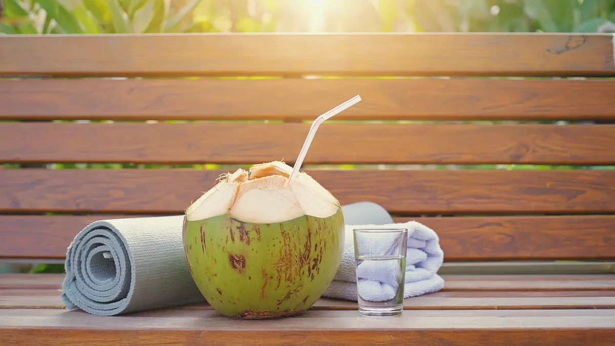 Eau de coco : 5 bienfaits réels et le comparatif essentiel 4 Noix de coco verte avec une paille, verre d'eau, tapis de yoga et serviette sur un banc après une séance